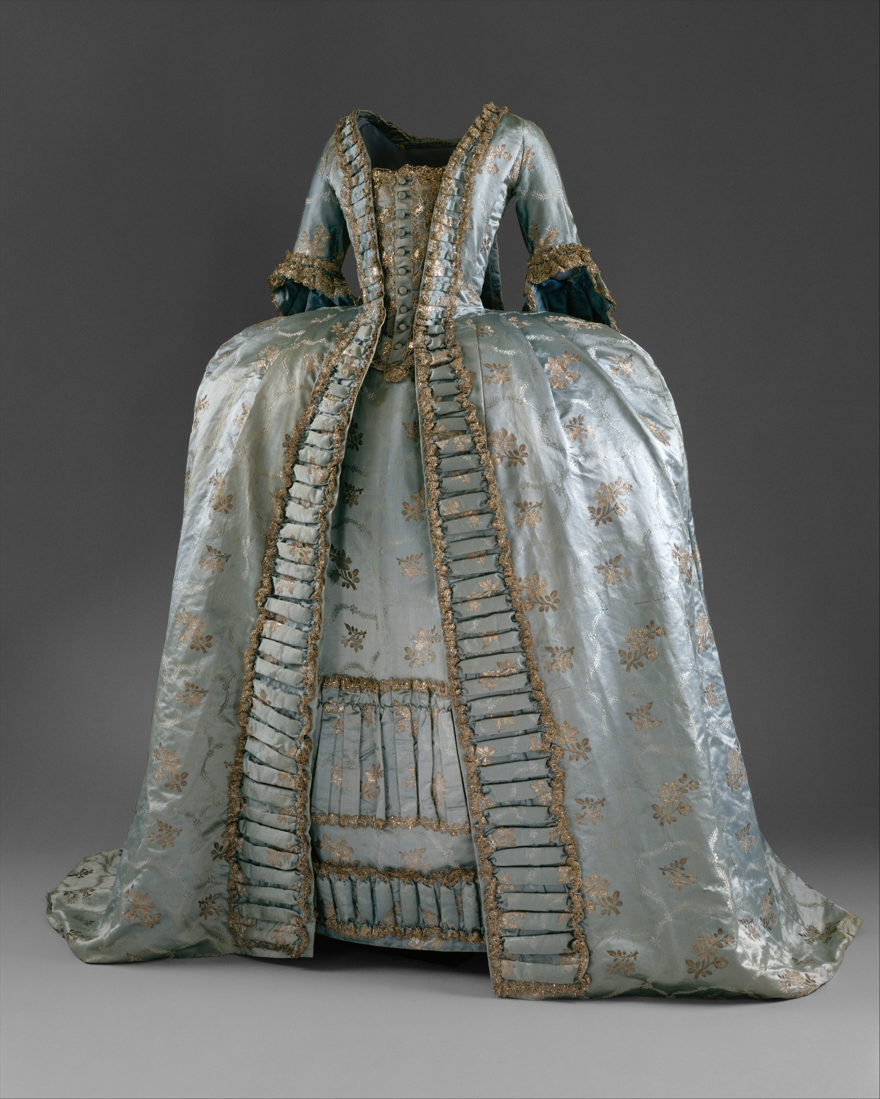 This image is of a robe a la francaise from the mid 1760s. It is a large, boxy dress made of light blue fabric and metallic embellishments, pleated fabric decorates the front of the dress from the neckline down the front of the skirt.