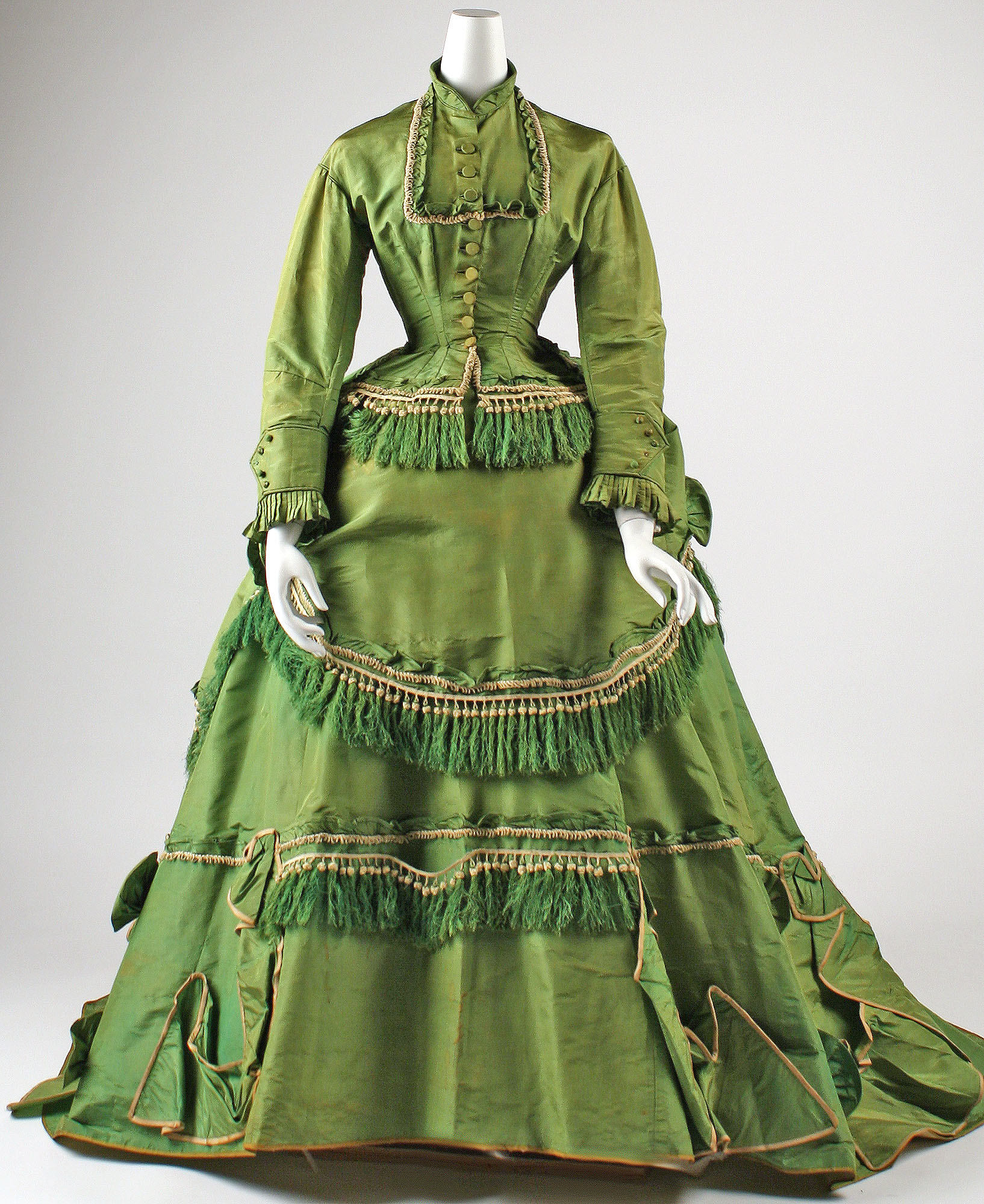 This is the image of a green, tasseled dress from 1868, and is American in origin. The dress is sharply tailored, with a narrow waist and voluminous skirt.