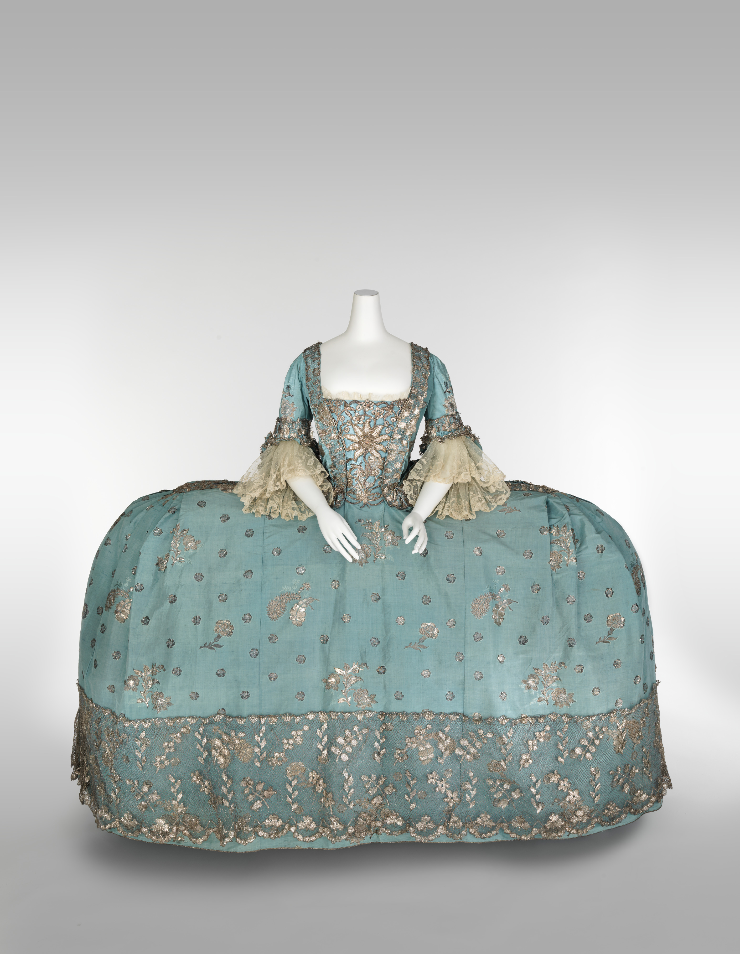 This is an image of an 1750 British court dress in teh style of the robe a la francaise; except in the extreme. The dress is almost twice as wide as the person wearing it, the light-blue fabric with metallic accents fitted over wide panniers.