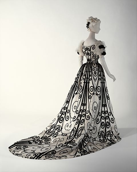 An image of a House of Worth evening gown from the end of the 19th century. The body of the dress is white, with intricate black flocking adorning the bodice, sleeves, skirt, and train.