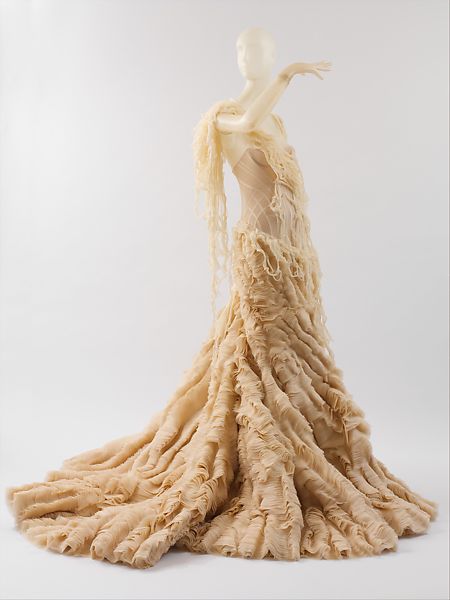 An image of Alexander McQueen's 2003 Oyster Dress from his Irere collection. The dress is a an a-line silhouette made of gauzy, mottled off-white fabric The dress appears ragged, as through it were composed of layers of torn gauze.